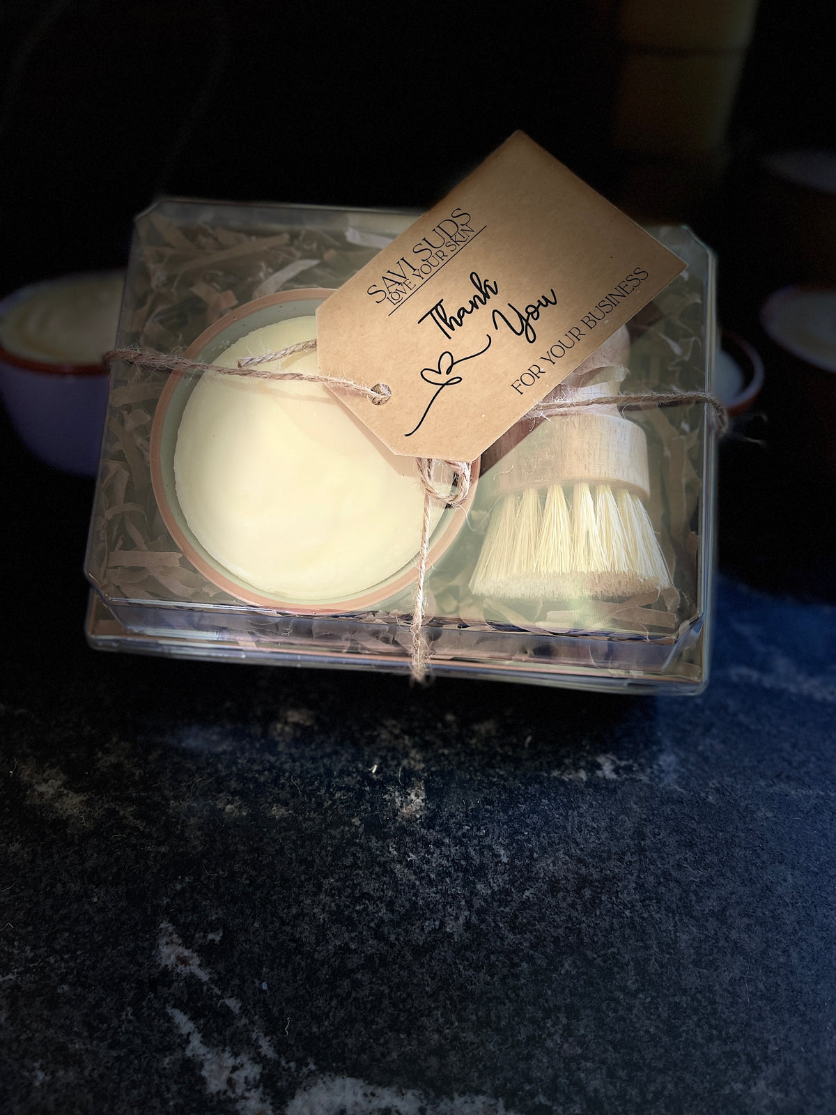 A boxed gift set of solid dish soap in a ramekin and a bamboo dish brush. Packaged in a box with shredded paper around it, and a clear top. A twine string is tied around it with a kraft cardstock tag