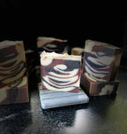 Handcrafted marbled soap bars with swirls of cream, brown, and dark, displayed on a dark reflective surface with one bar resting on a small gray stone stand.