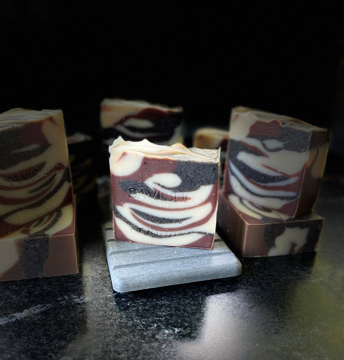 Handcrafted marbled soap bars with swirls of cream, brown, and dark, displayed on a dark reflective surface with one bar resting on a small gray stone stand.