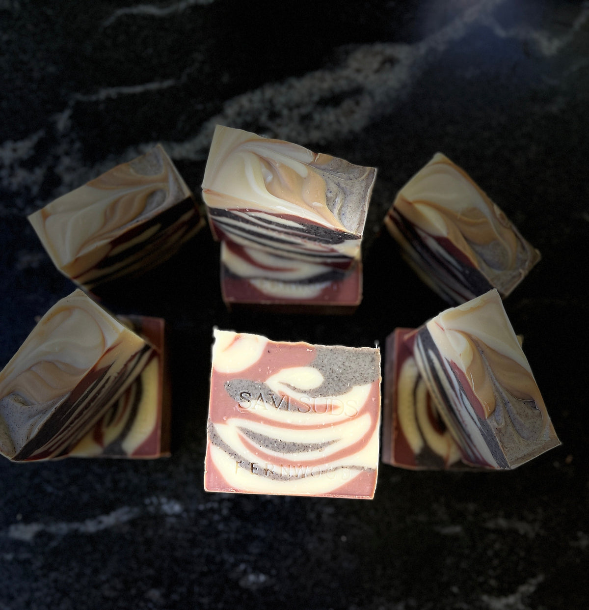Several artisanal marbled soap bars arranged on a dark marble surface, with one soap bar centered and facing forward to show the label. The soaps feature tiger swirls of cream, brown, and dark gray.