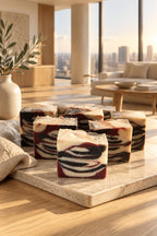 Several rectangular bars of marbled artisanal soap are arranged on a light surface in a light filled room. The front soap bar is in sharp focus, showing a tiger swirl pattern of cream, dark grey, and reddish-brown, while the soap bars behind it are softly blurred.