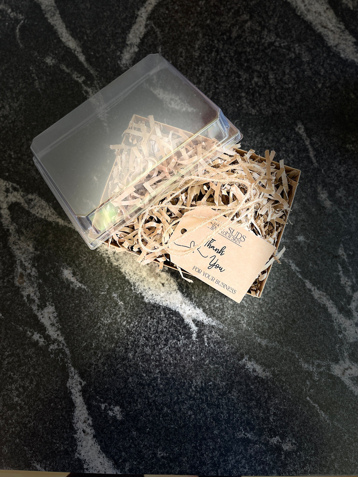 A clear plastic gift box with a transparent lid, partially open and filled with shredded kraft paper and a small beige tag, placed on a dark marbled surface.