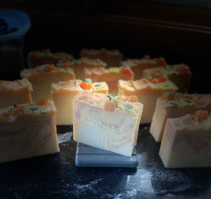 A handmade soap bar labeled 'SAVI SUDS', standing upright on a small slate block. The soap has cream, light orange, and rust swirls and is topped with soap dough pumpkins and sunflowers, sprinkled with gold mica. The soap is surrounded by other similar soaps on a dark, marbled surface
