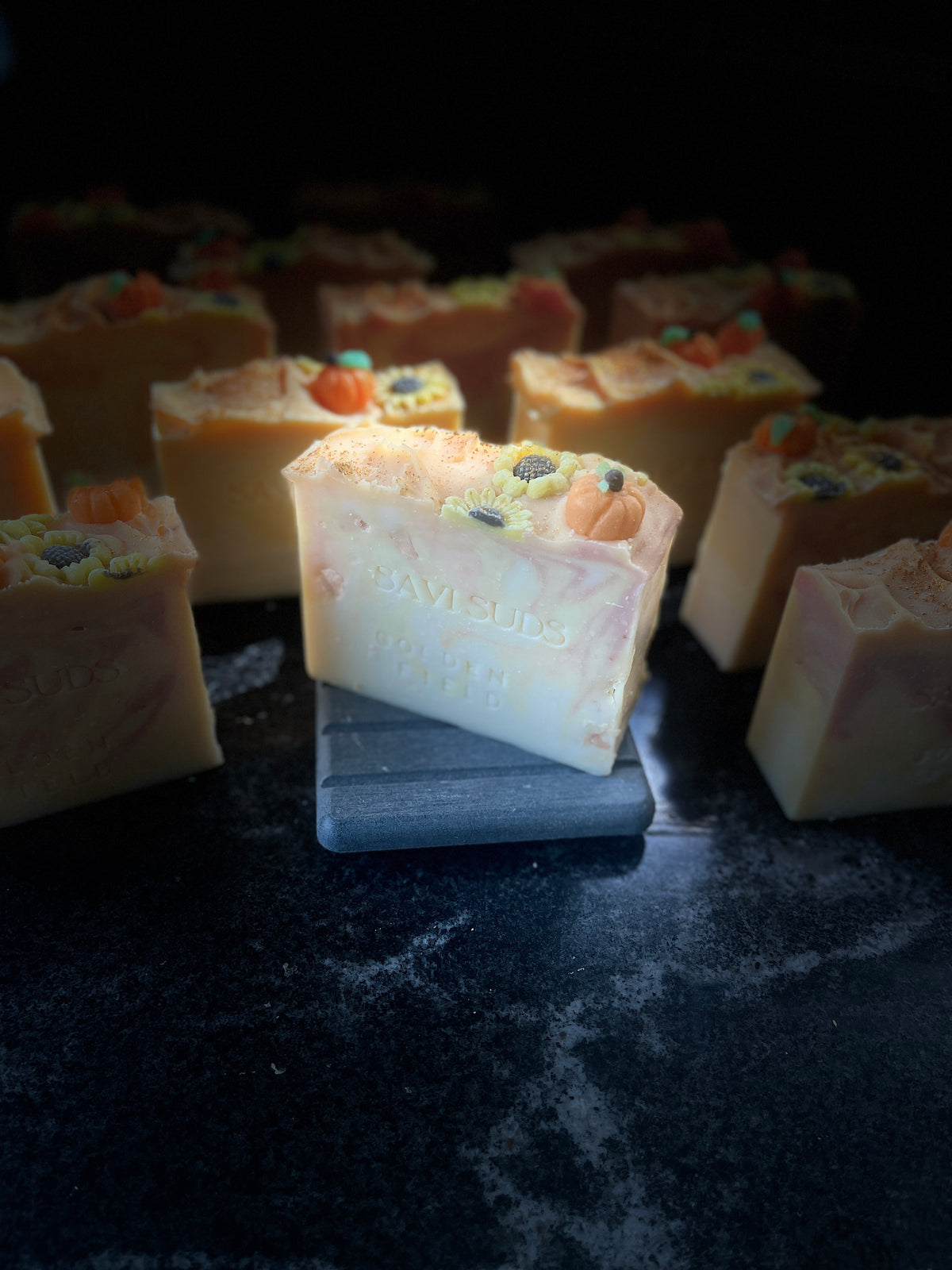 A handmade soap bar labeled 'Savi Suds' sits on a small dark slate block, surrounded by other soap bars in a dimly lit setting.