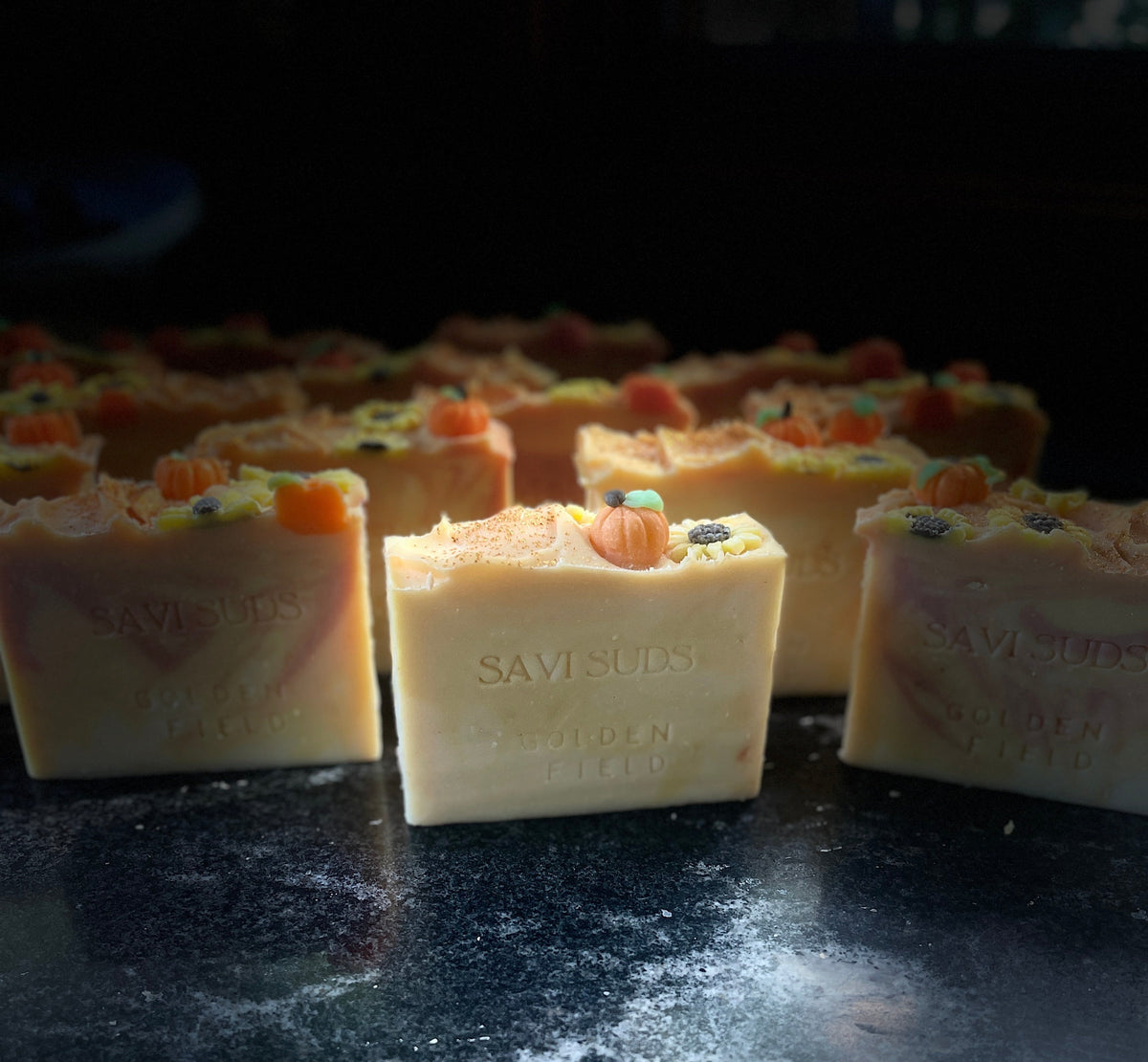 Several handmade soap bars with a cream-colored base and soap dough pumpkins and sunflowers on top, arranged on a dark, textured surface.