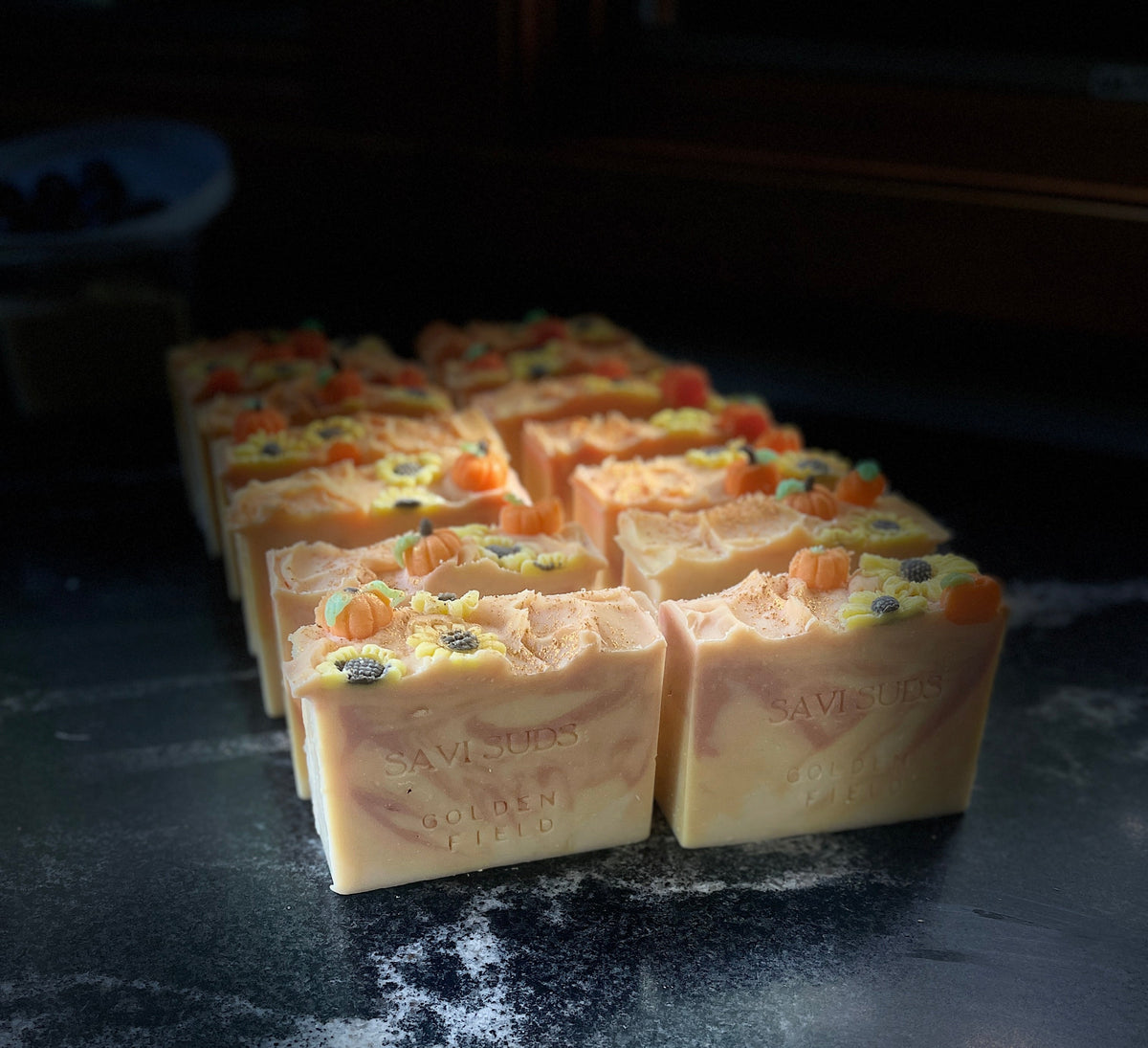 A row of handmade rectangular soap bars with swirled cream and orange tones and small pumpkins and sunflowers made of soap dough on top, arranged on a dark stone surface