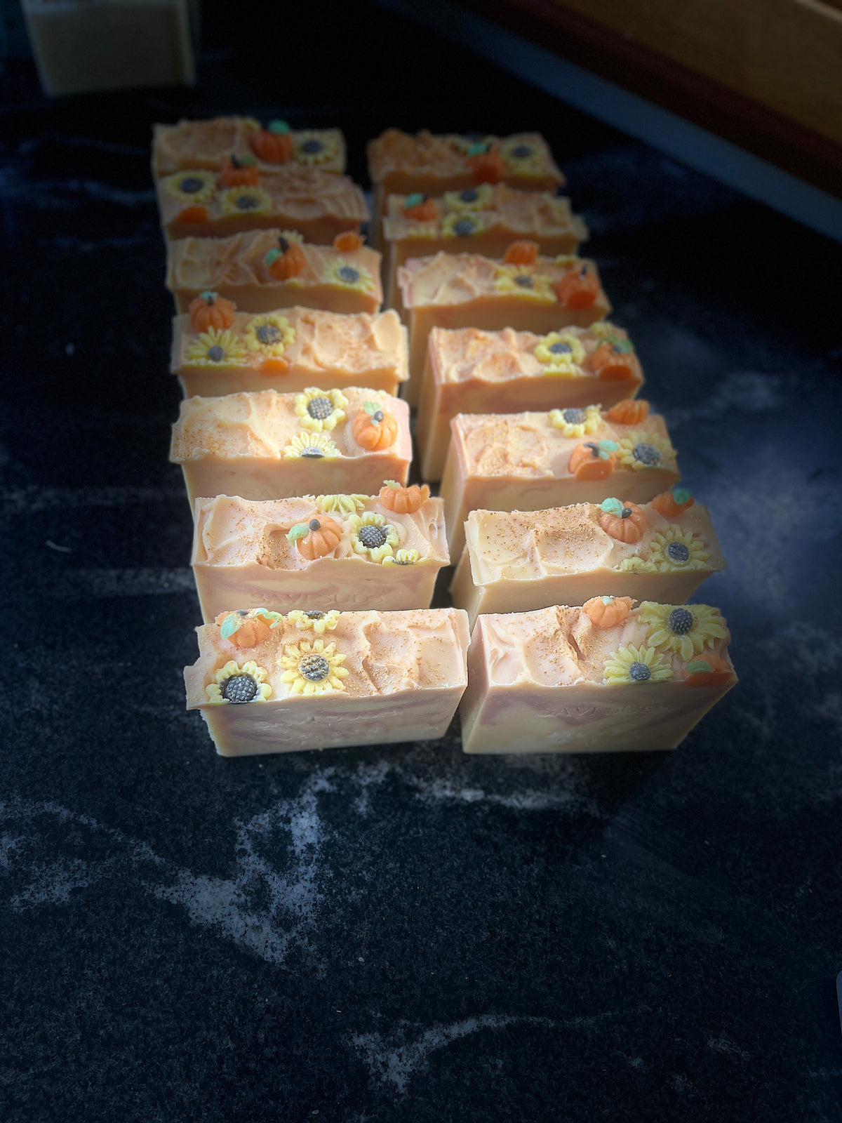 A set of rectangular handmade soap bars decorated with orange pumpkins and yellow sunflowers made of soap dough, arranged on a dark textured surface.