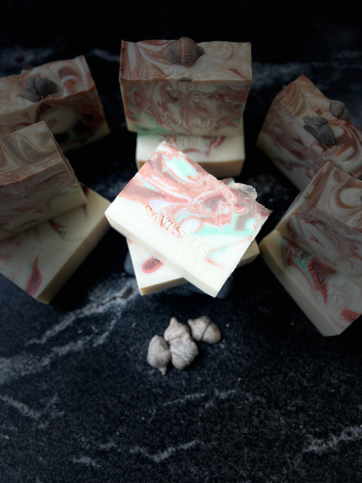 Several artisanal soap bars with marbled pink, cream, and mint green swirls are arranged on a dark textured surface. One soap bar in the center is slightly tilted and shows the brand name 'SAVI SUDS' stamped on it.