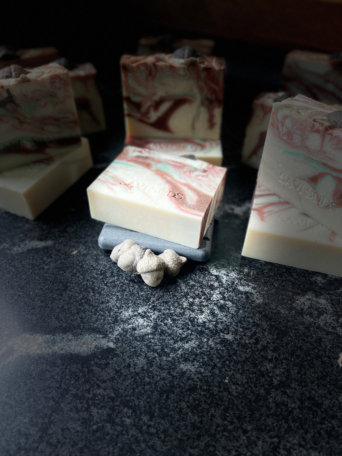 Handcrafted marbled soap bars with cream, green, and brown swirls, styled on a dark textured surface with small acorns made of soap dough in front