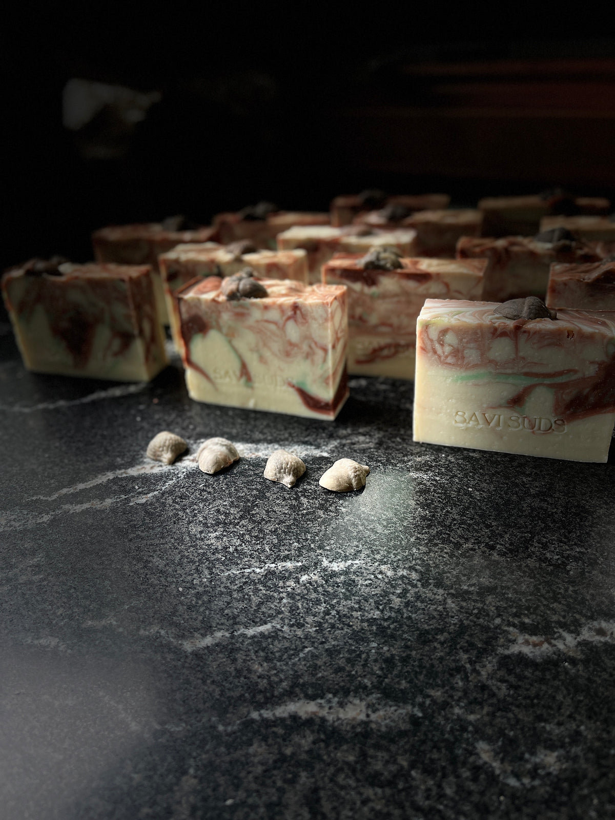 Handcrafted rectangular soap bars with a cream base and swirling brown and green marbled patterns, arranged on a dark stone surface with small acorns in the foreground.