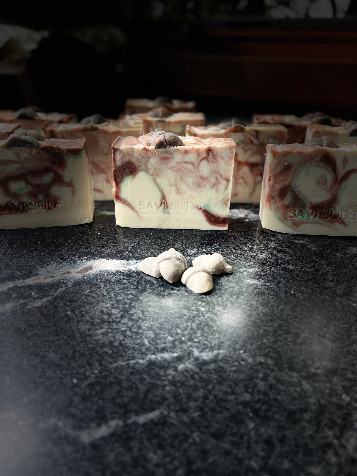 Several rectangular bars of handmade soap with a cream base and dark reddish-brown and green swirls are arranged on a dark marble-like surface. Small acorn embed pieces are placed on top of the bars, and a few loose pieces sit in front.