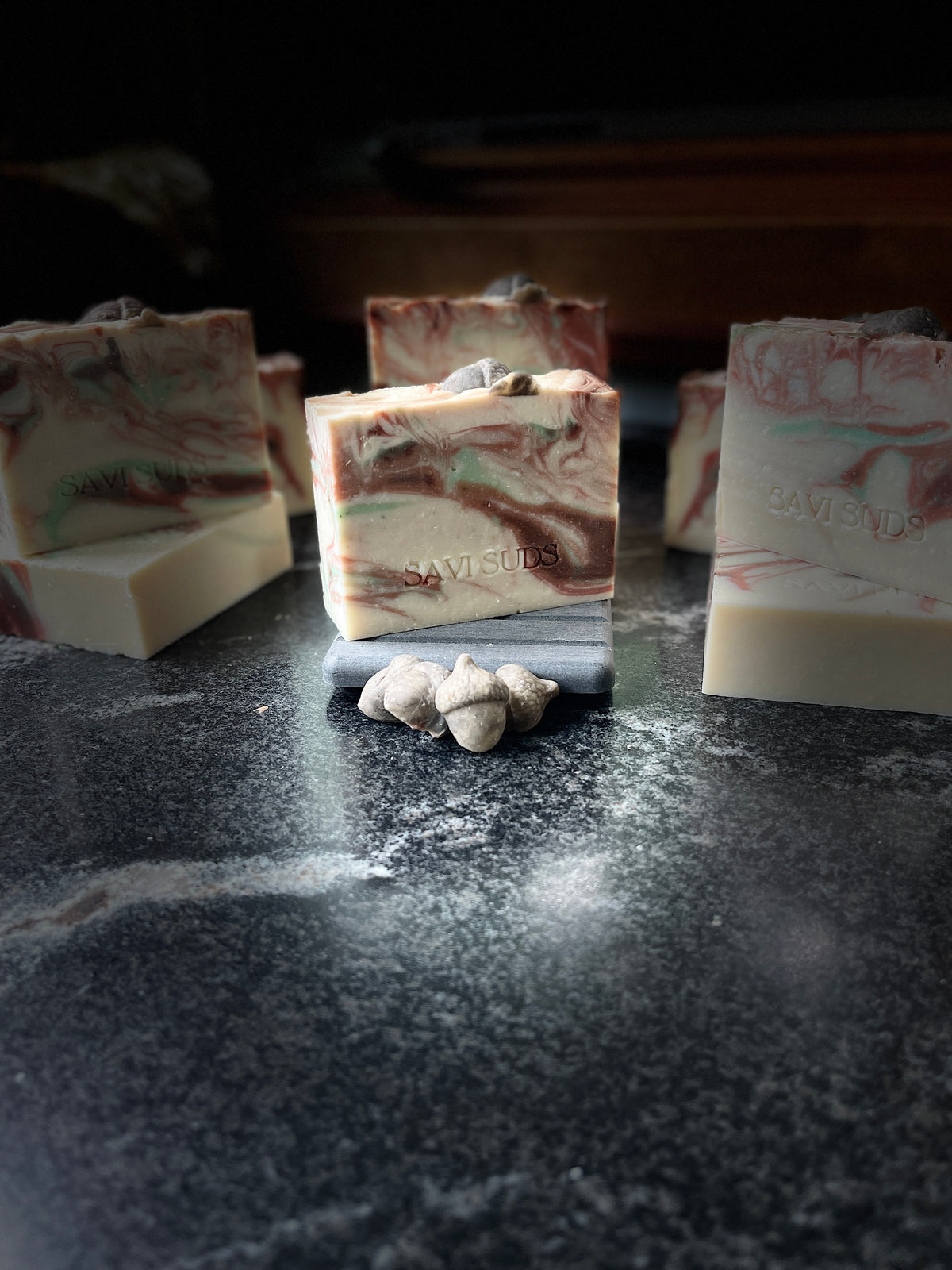 Several rectangular bars of marbled soap are arranged on a dark stone surface, with one bar centered on a small gray block and a few small acorn shaped soap dough pieces in front. The soap has an off-white base with brown and green marbling, and the center bar appears stamped with the brand name SAVI SUDS.
