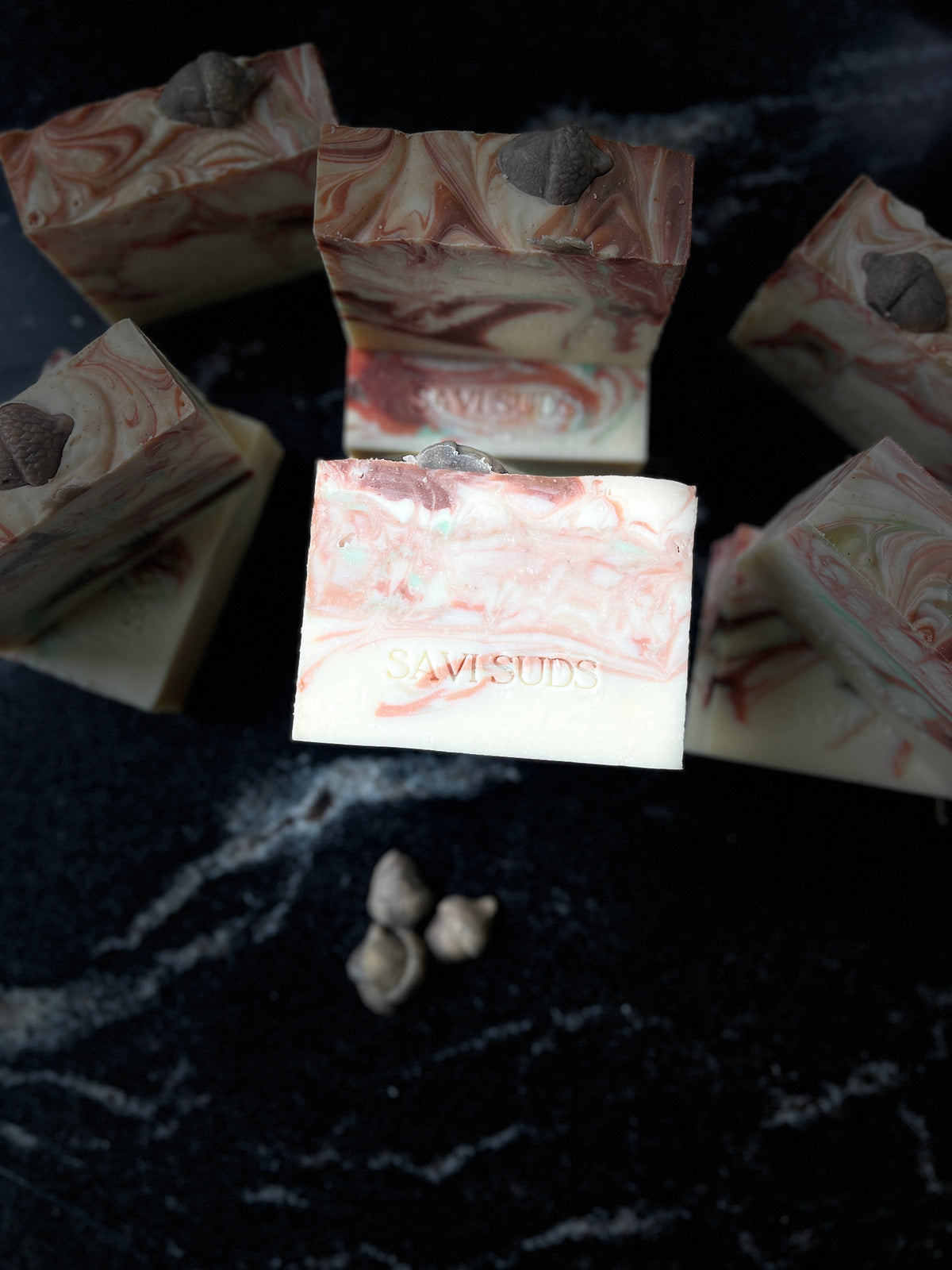 Several handmade marbled soaps with cream, green, and brown swirls are arranged on a dark marble surface. One soap in the foreground is centered and reads 'Savi Suds' on the front, with small acorns made of soap dough placed near the base.