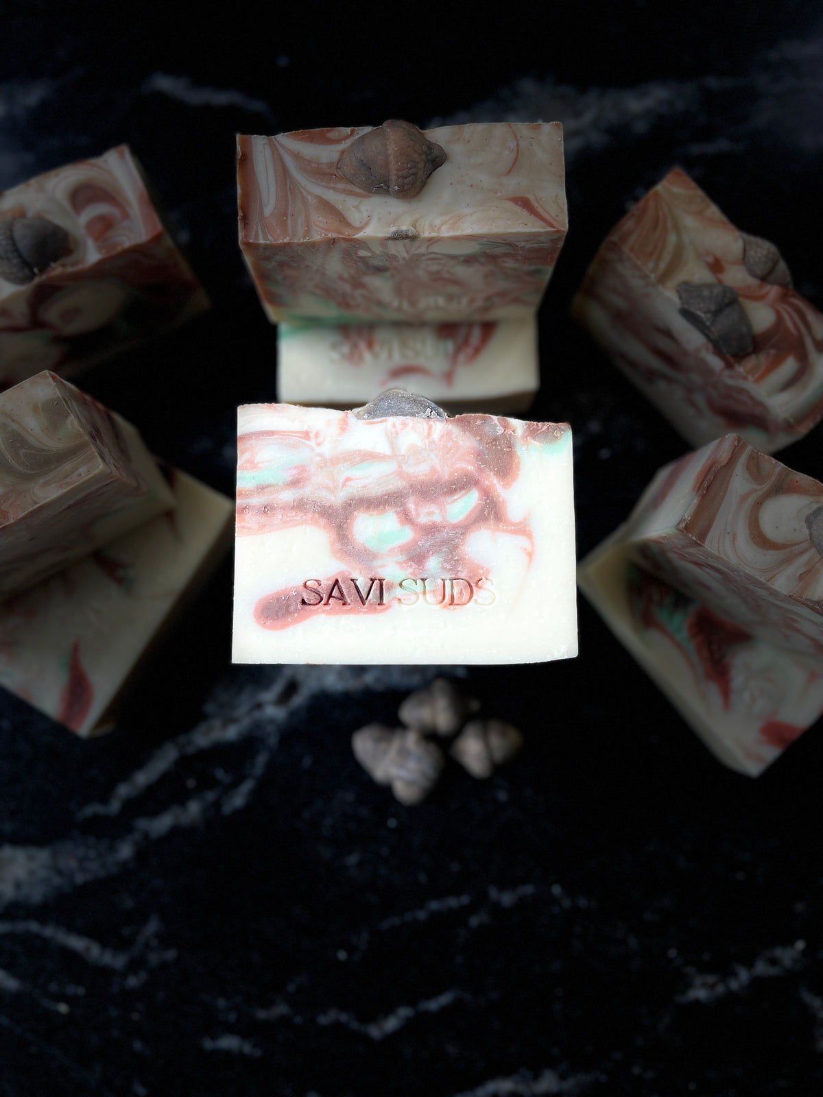 Five artisanal marbled soap bars with acorn inclusions are arranged on a dark, textured background. The central soap bar is upright and clearly displays the brand name 'SAVISUDS' stamped on the front, while the other bars are positioned around it.