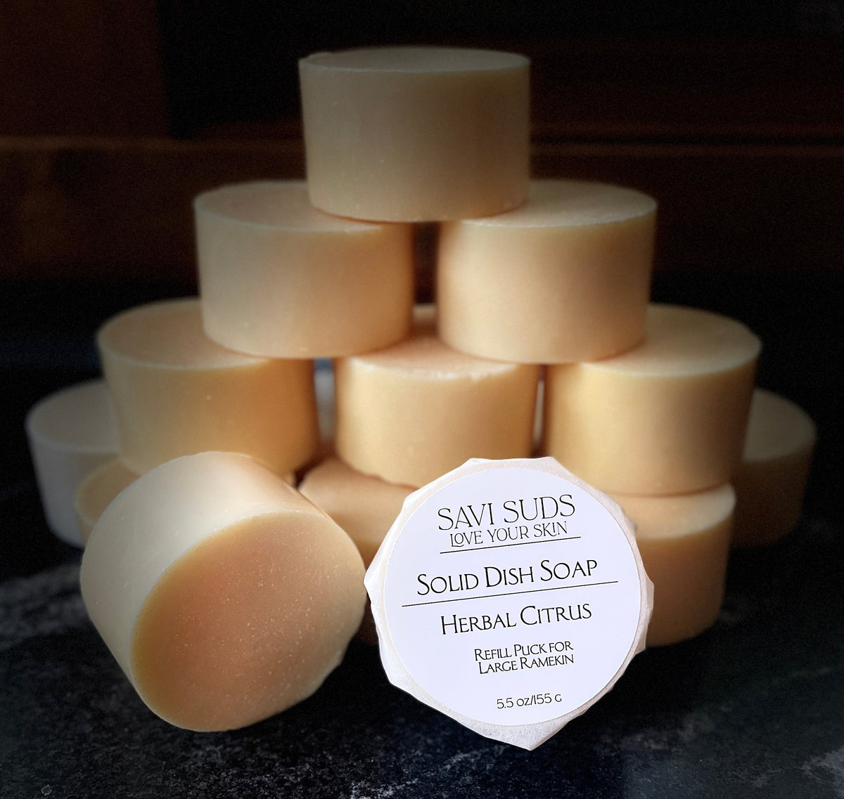 A stack of solid dish soap refill bars with one in front wrapped in bleach free coffee filter that reads 'SAVI SUDS' and 'SOLID DISH SOAP HERBAL CITRUS', set on a dark textured countertop.