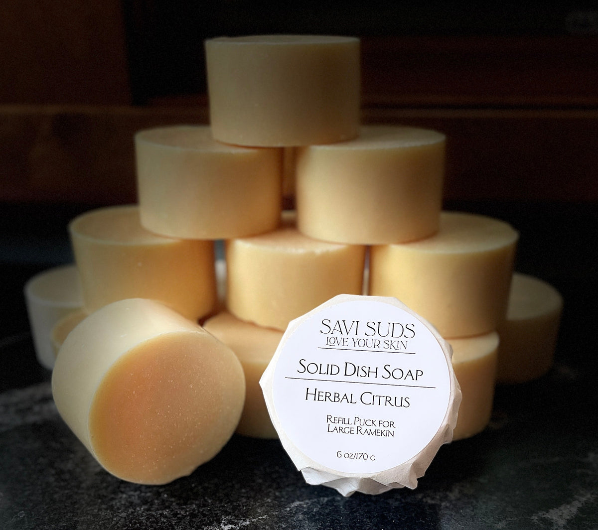 A stack of solid dish soap refill bars with one in front wrapped in bleach free coffee filter that reads 'SAVI SUDS' and 'SOLID DISH SOAP HERBAL CITRUS', set on a dark textured countertop.