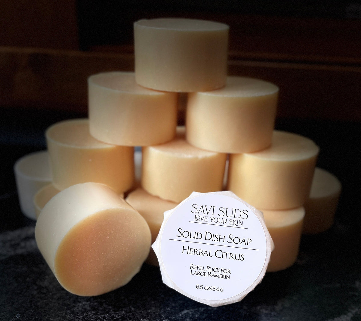 A stack of solid dish soap refill bars with one in front wrapped in bleach free coffee filter that reads 'SAVI SUDS' and 'SOLID DISH SOAP HERBAL CITRUS', set on a dark textured countertop.