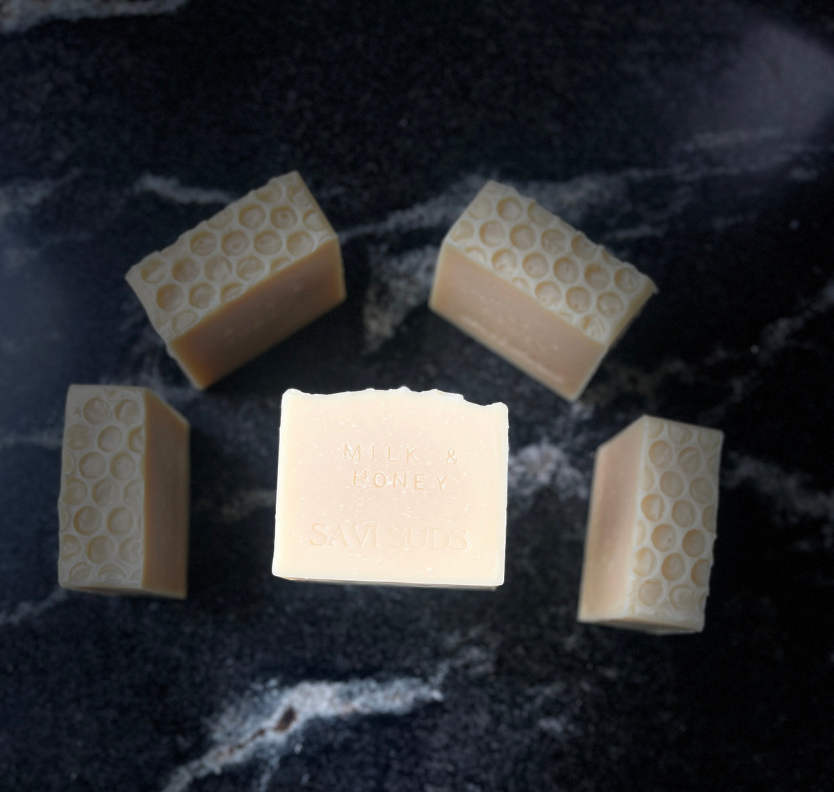 Five bars of handmade soap arranged on a dark marble surface, with one central bar and four surrounding bars. The soaps are a warm cream color with a honeycomb texture on the top