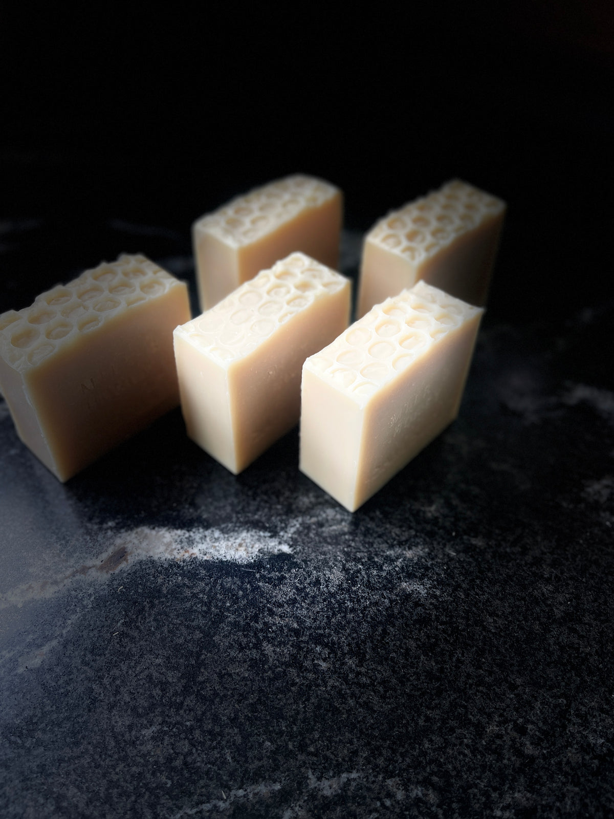 Five square bars of handmade soap with a honeycomb pattern on top, shown on a dark textured surface against a black background.
