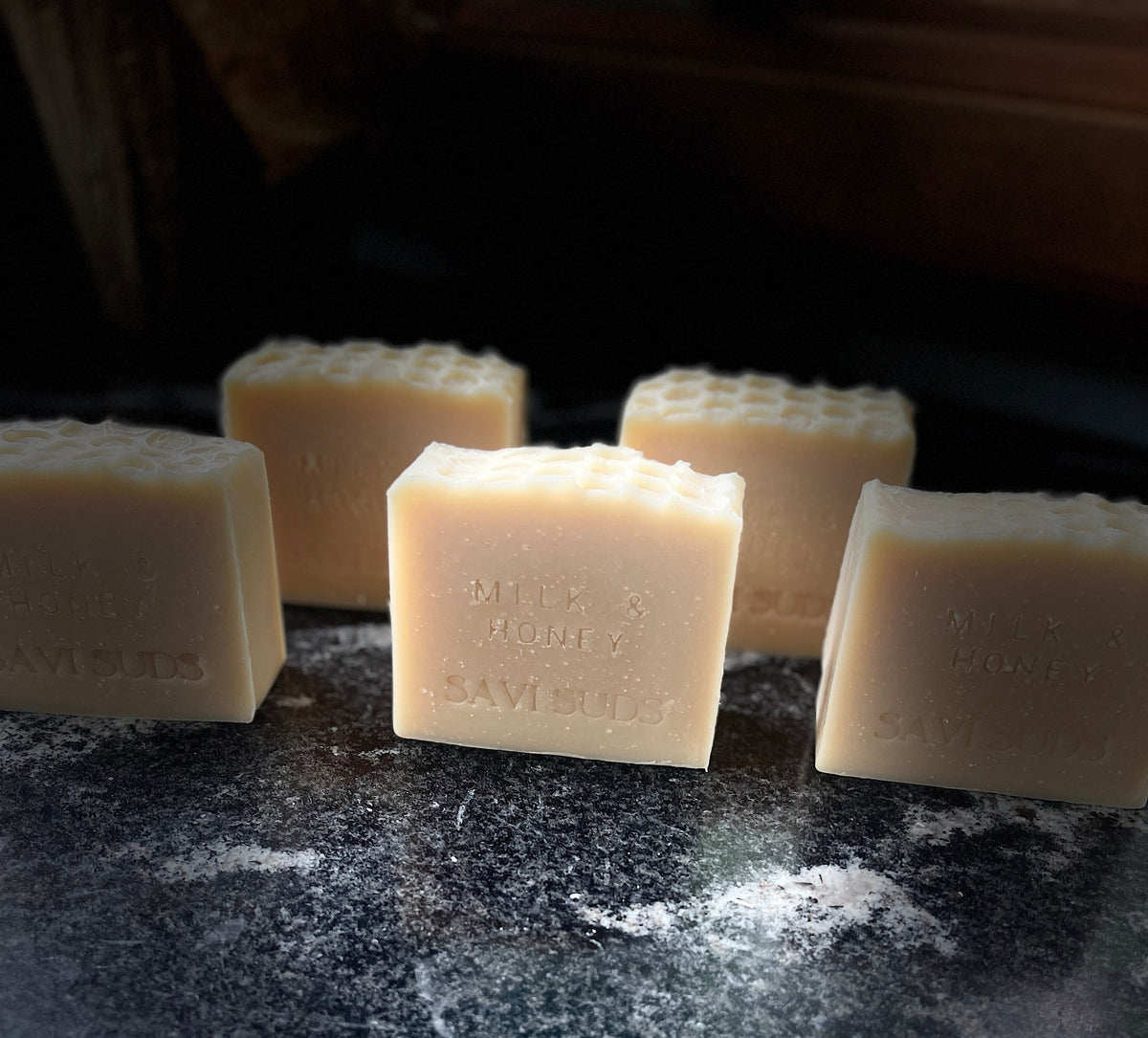Five square bars of handmade soap with a honeycomb pattern on top, shown on a dark textured surface against a black background.
