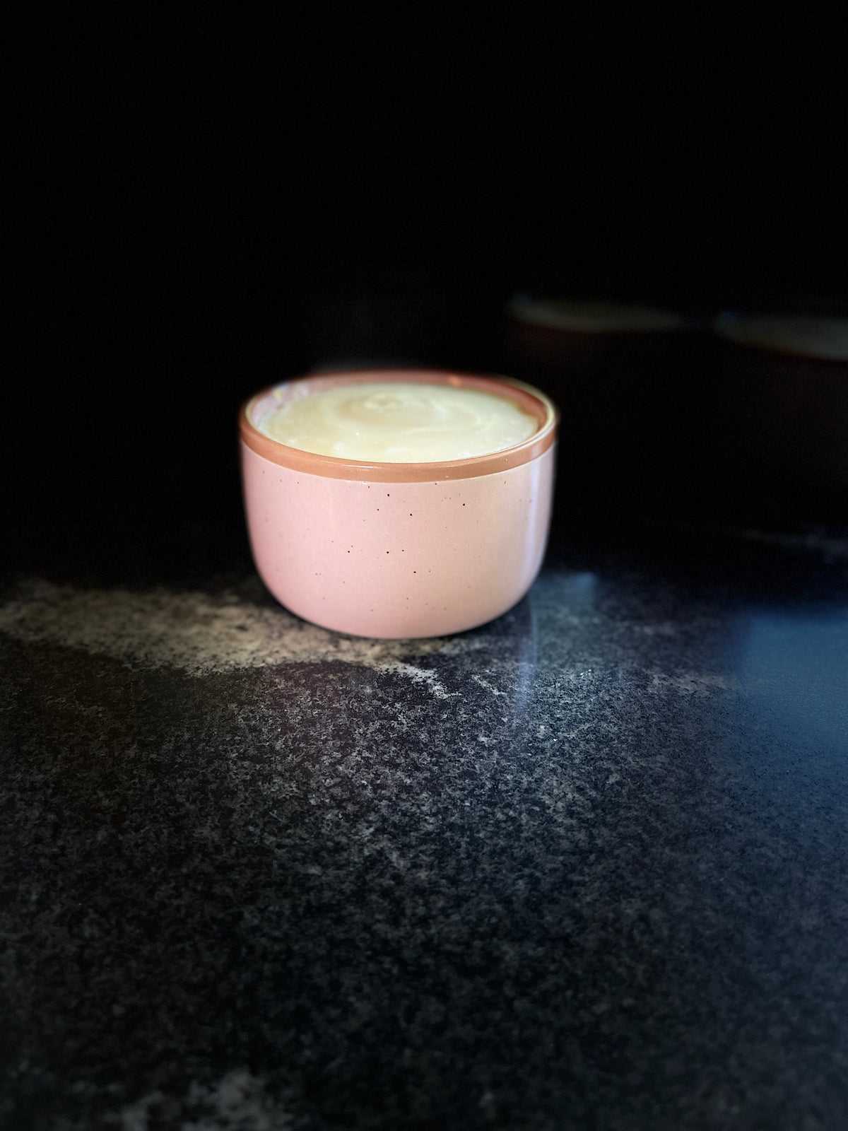A speckled pink ceramic bowl with a warm brown rim, filled with solid dish soap set on a dark textured surface against a black background.