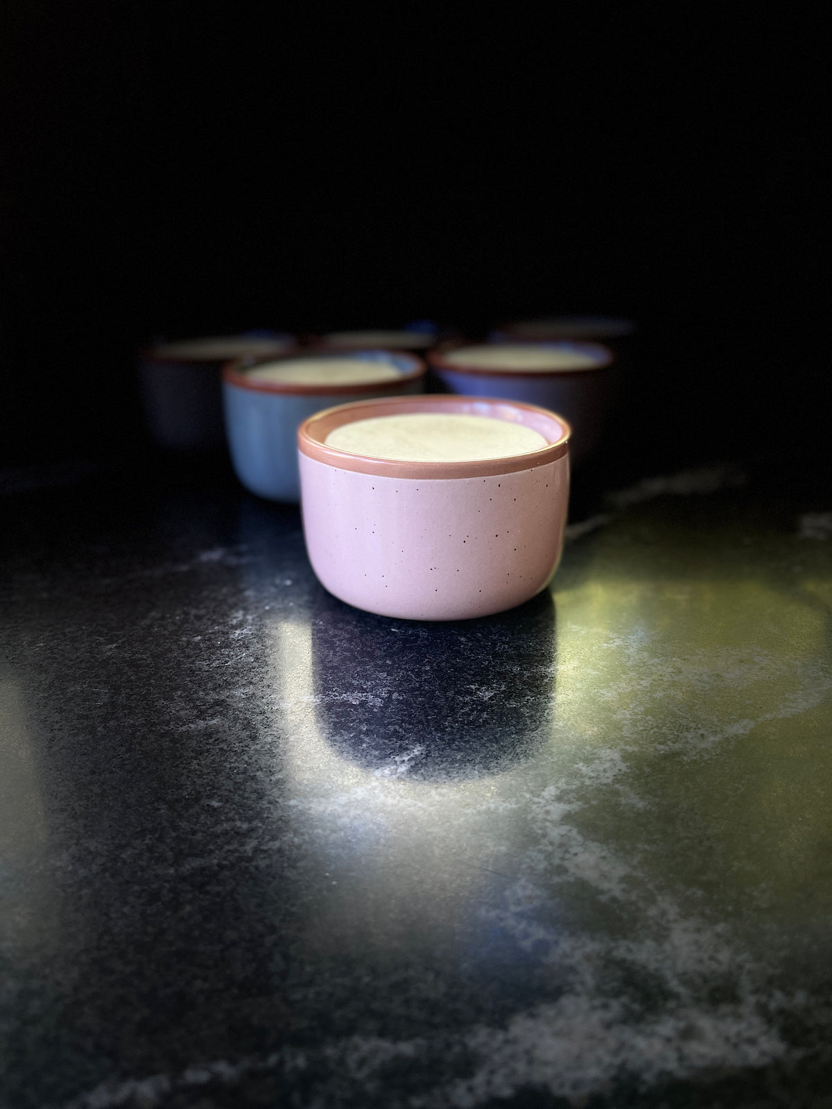 A speckled ceramic bowl with a warm brown rim, filled with solid dish soap set on a dark textured surface against a black background.