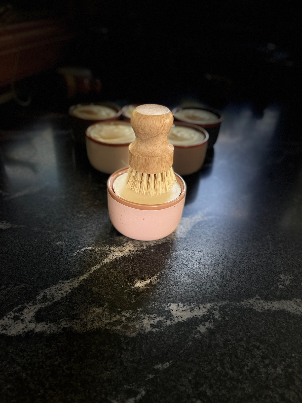 A large blush pink-colored ceramic container filled with solid dish soap, topped with a wooden-handled brush with natural bristles, set on a dark marble-like surface with several similar containers blurred in the background.