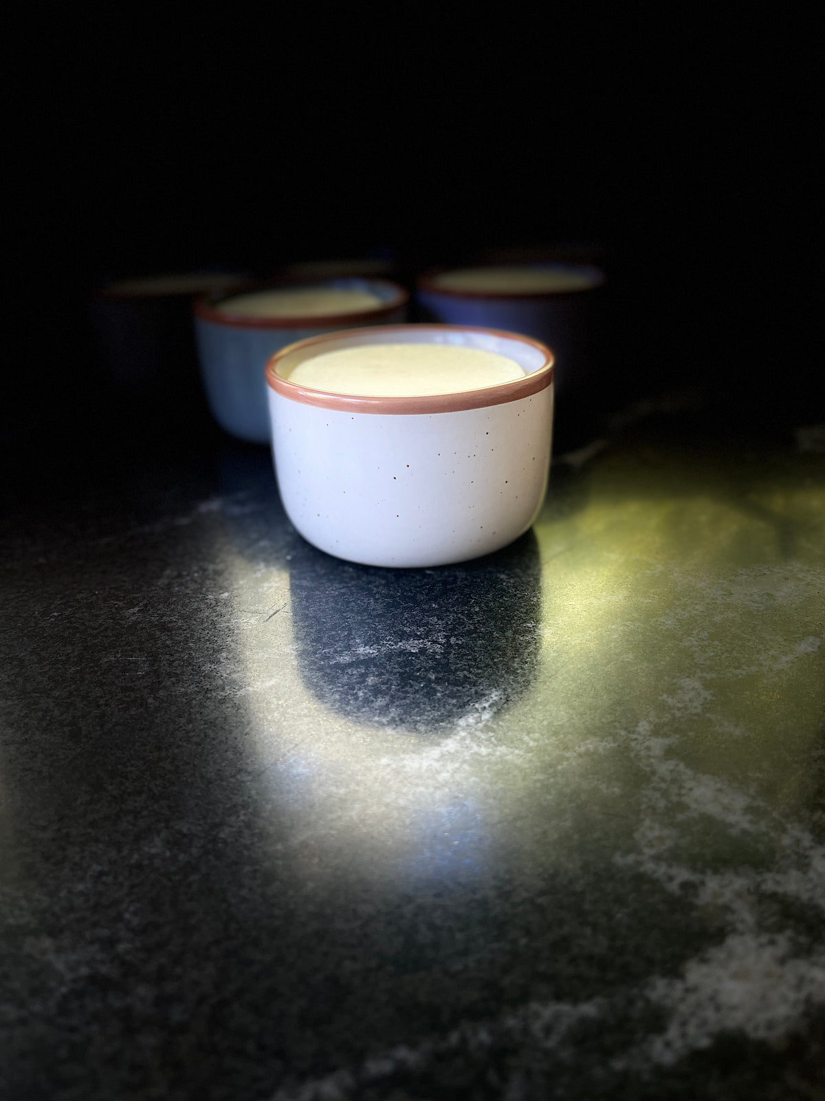 A speckled ceramic bowl with a warm brown rim, filled with solid dish soap set on a dark textured surface against a black background.