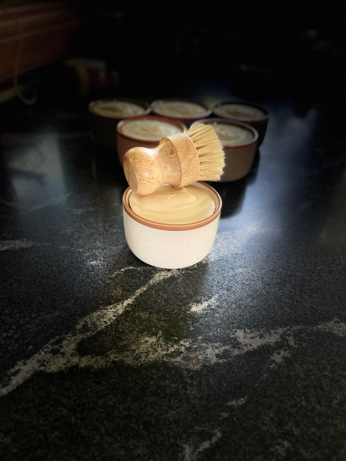 A large cream-colored ceramic container filled with solid dish soap, topped with a wooden-handled brush with natural bristles, set on a dark marble-like surface with several similar containers blurred in the background.