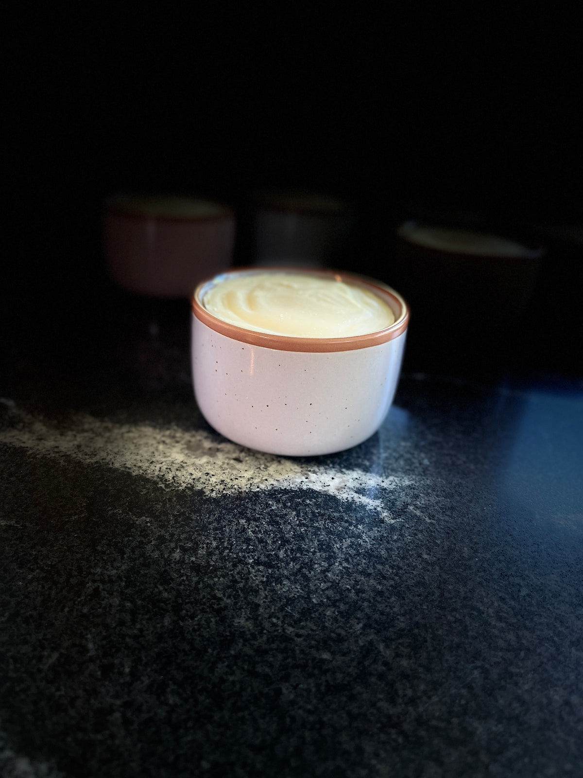 A speckled lavender ceramic dish with warm brown rim, filled with solid dish soap, sitting on a dark textured surface 