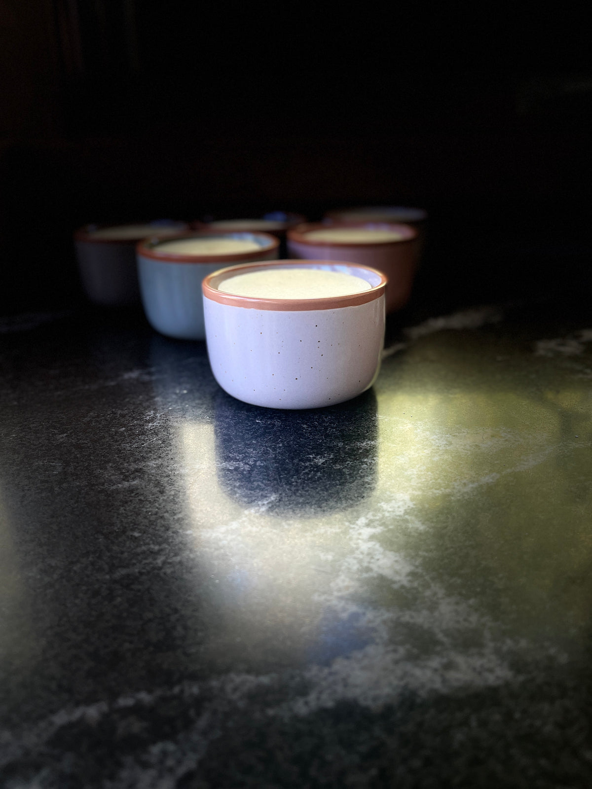 A speckled ceramic bowl with a warm brown rim, filled with solid dish soap set on a dark textured surface against a black background.