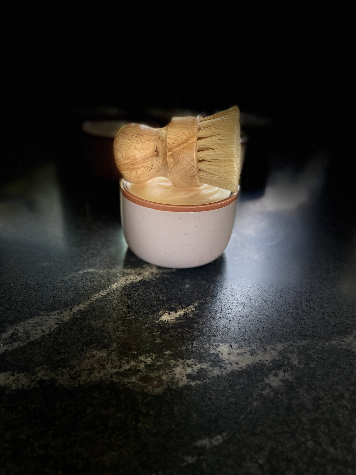 A large pale lavender-colored ceramic container filled with solid dish soap, topped with a wooden-handled brush with natural bristles, set on a dark marble-like surface with several similar containers blurred in the background.