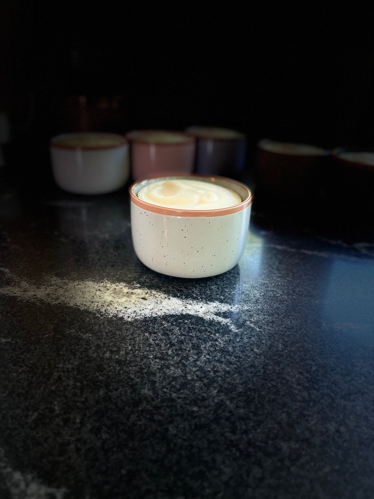 A speckled sage ceramic cup with a warm brown rim and solid dish soap inside, set on a dark countertop with other similar cups blurred in the background.