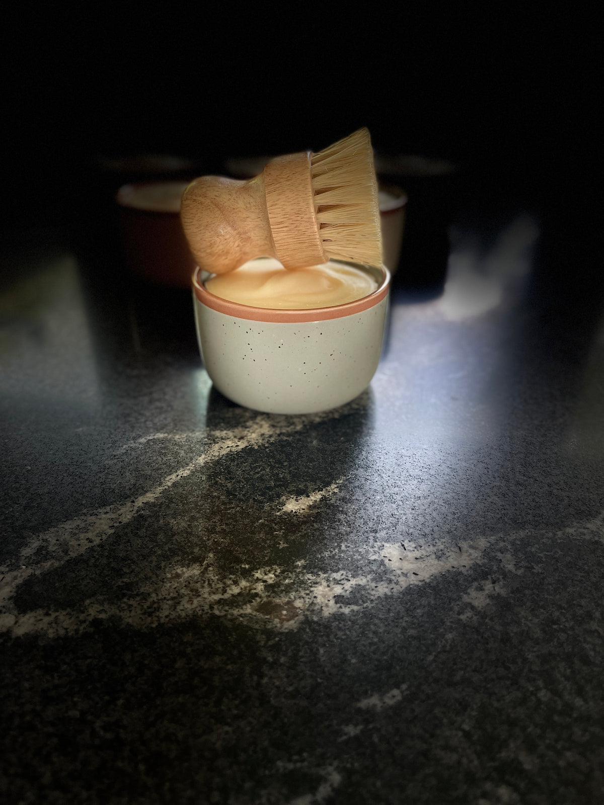 A large sage green-colored ceramic container filled with solid dish soap, topped with a wooden-handled brush with natural bristles, set on a dark marble-like surface with several similar containers blurred in the background.