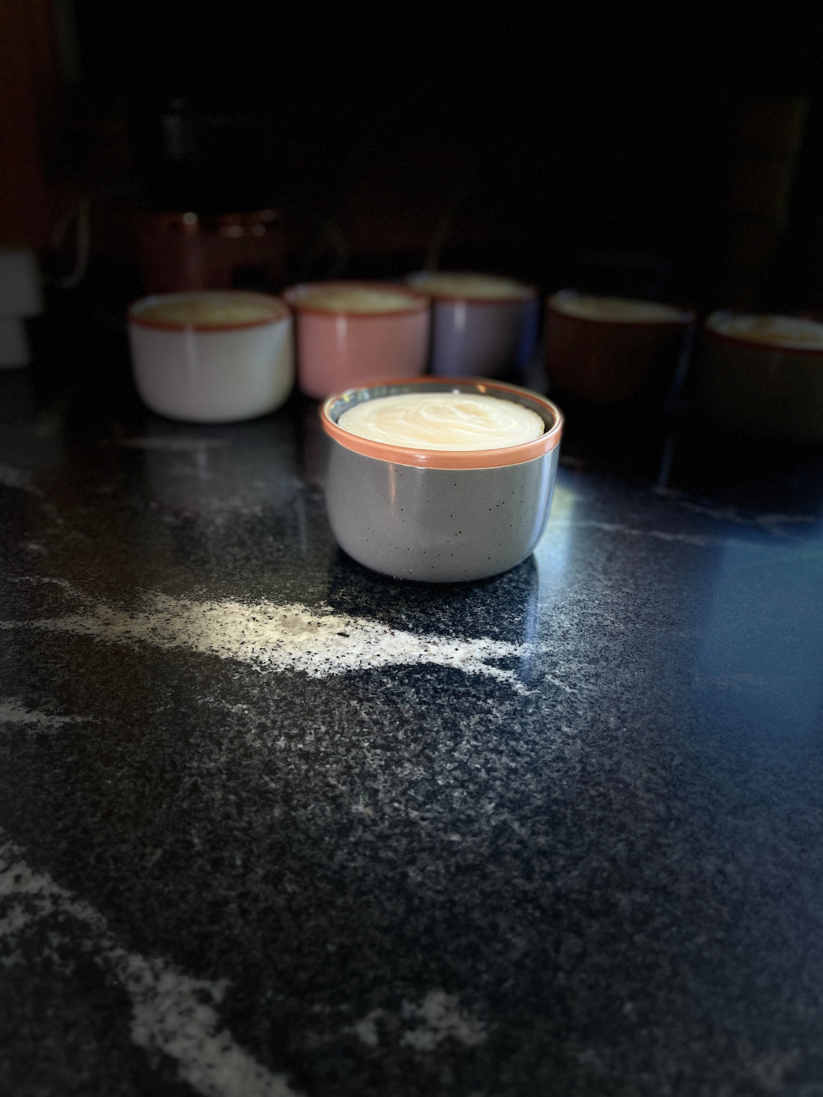 A speckled slate gray ceramic ramekin filled with solid dish soap, set on a dark countertop with several similar ramekins blurred in the background.