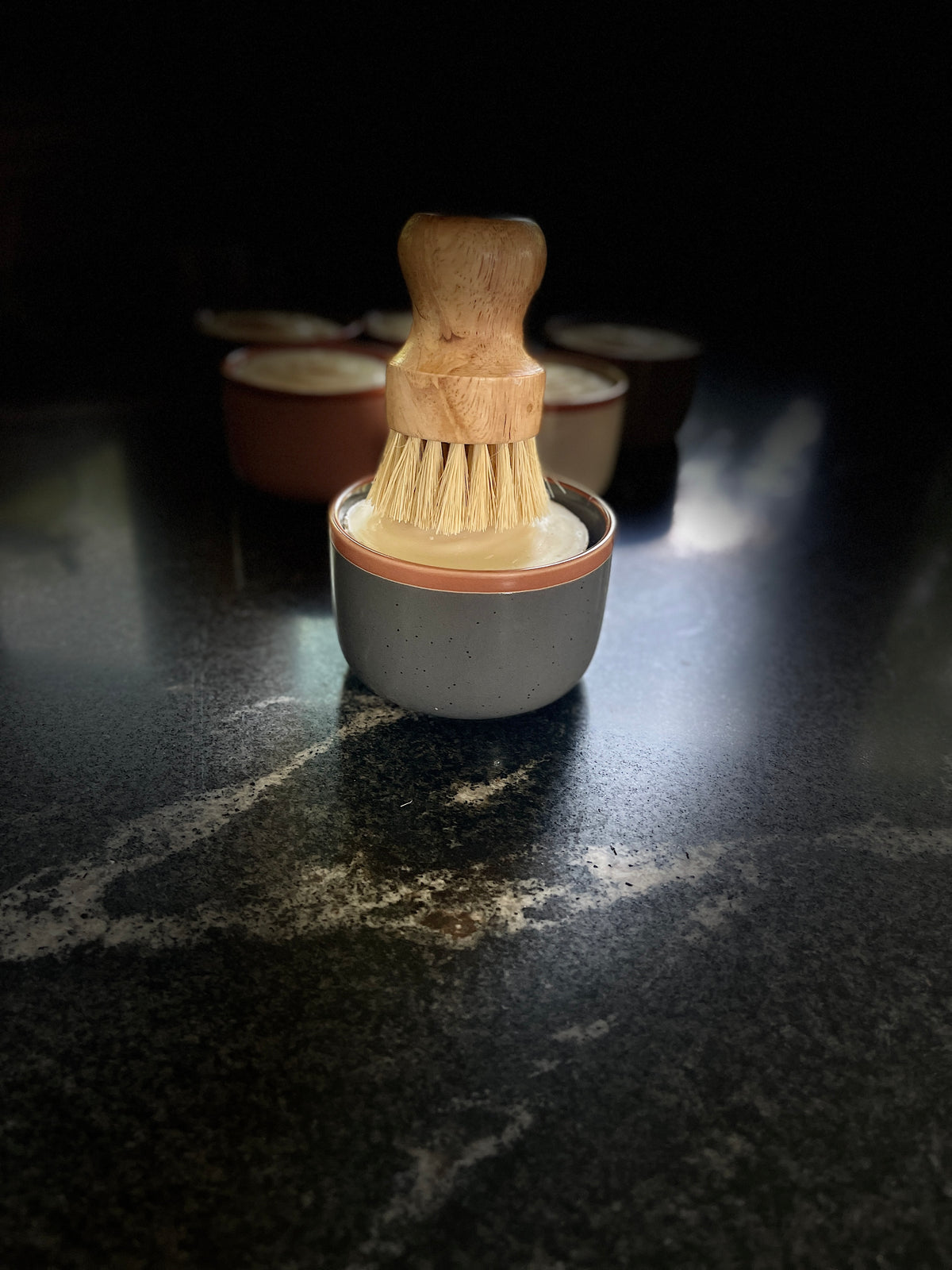 A large slate gray-colored ceramic container filled with solid dish soap, topped with a wooden-handled brush with natural bristles, set on a dark marble-like surface with several similar containers blurred in the background.