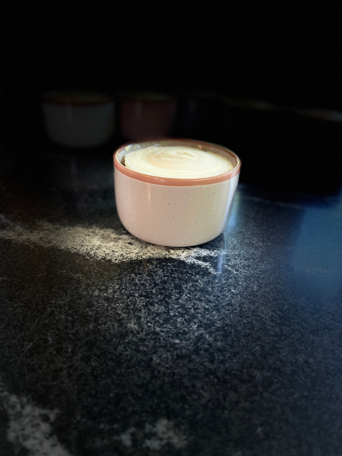 A speckled beige ceramic dish with a terracotta brown rim filled with solid dish soap, sitting on a dark, textured countertop with a soft glow reflecting off its surface.
