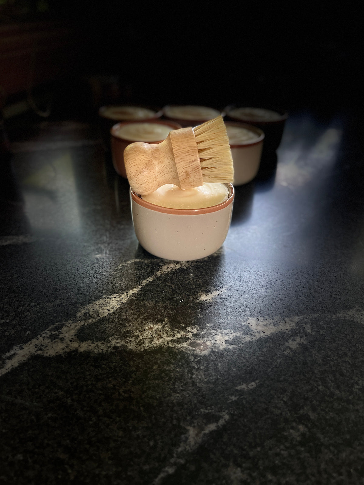 A large warm beige-colored ceramic container filled with solid dish soap, topped with a wooden-handled brush with natural bristles, set on a dark marble-like surface with several similar containers blurred in the background.