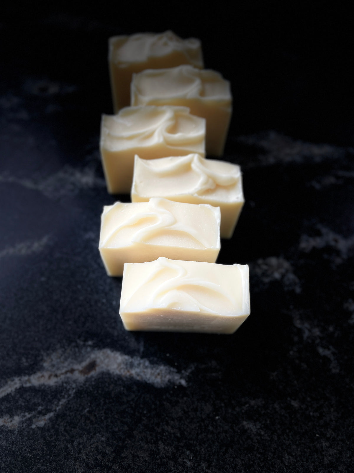 Six square bars of creamy off-white soap arranged in a diagonal row on a dark, textured surface.