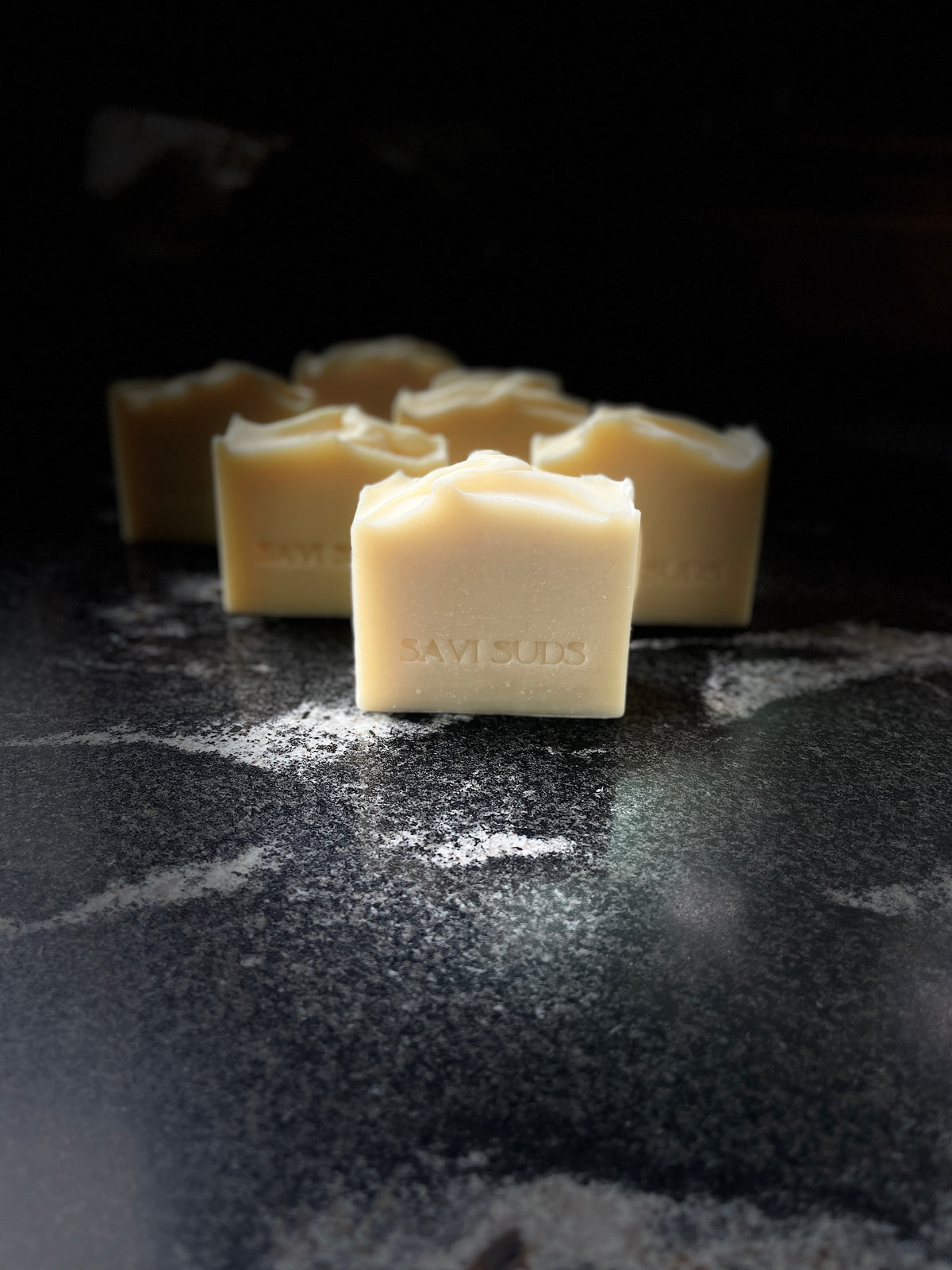 Six square bars of handmade cream-colored soap, with the front of the bars stamped with the brand name 'SAVI SUDS', arranged on a dark, textured surface 