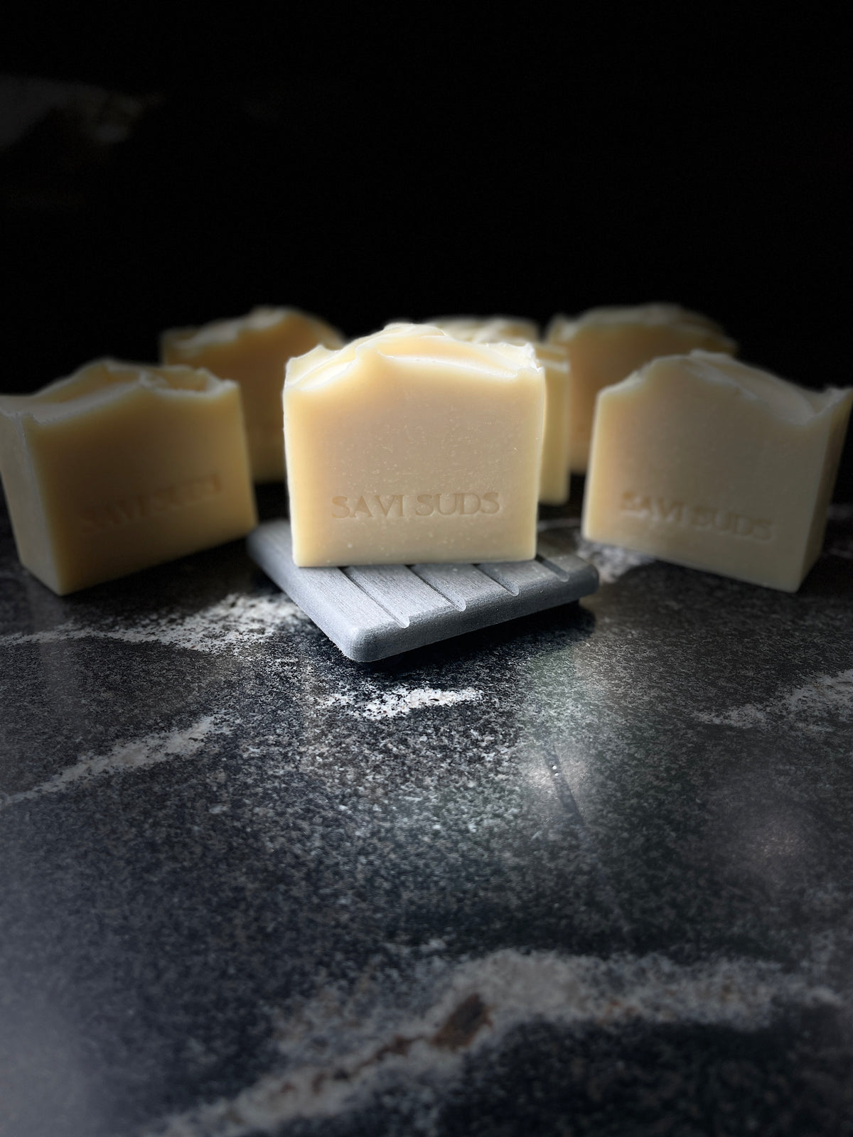 Six square bars of handmade cream-colored soap with 'SAVI SUDS' stamped on the front, arranged on a dark textured surface with one bar resting on a small gray block.