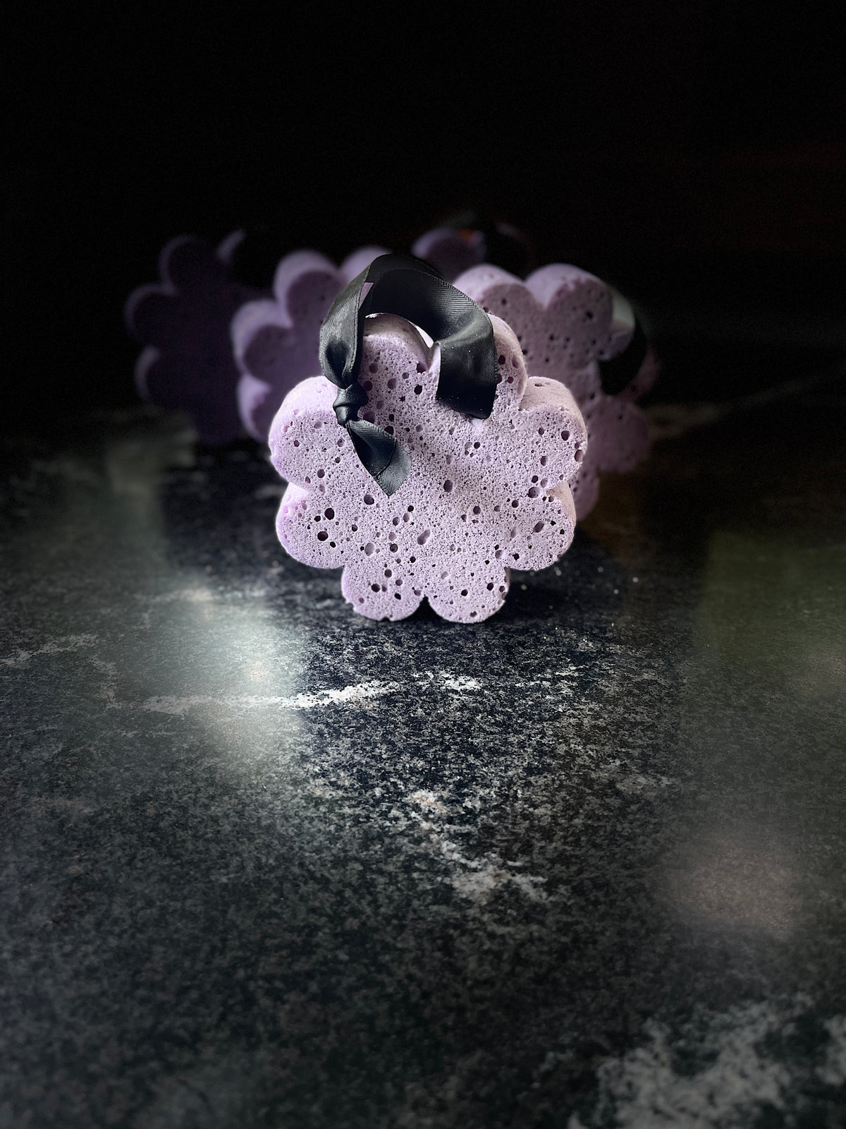 A set of purple flower-shaped bath and body sponge bars from Savi Suds, resting on a dark marble surface.