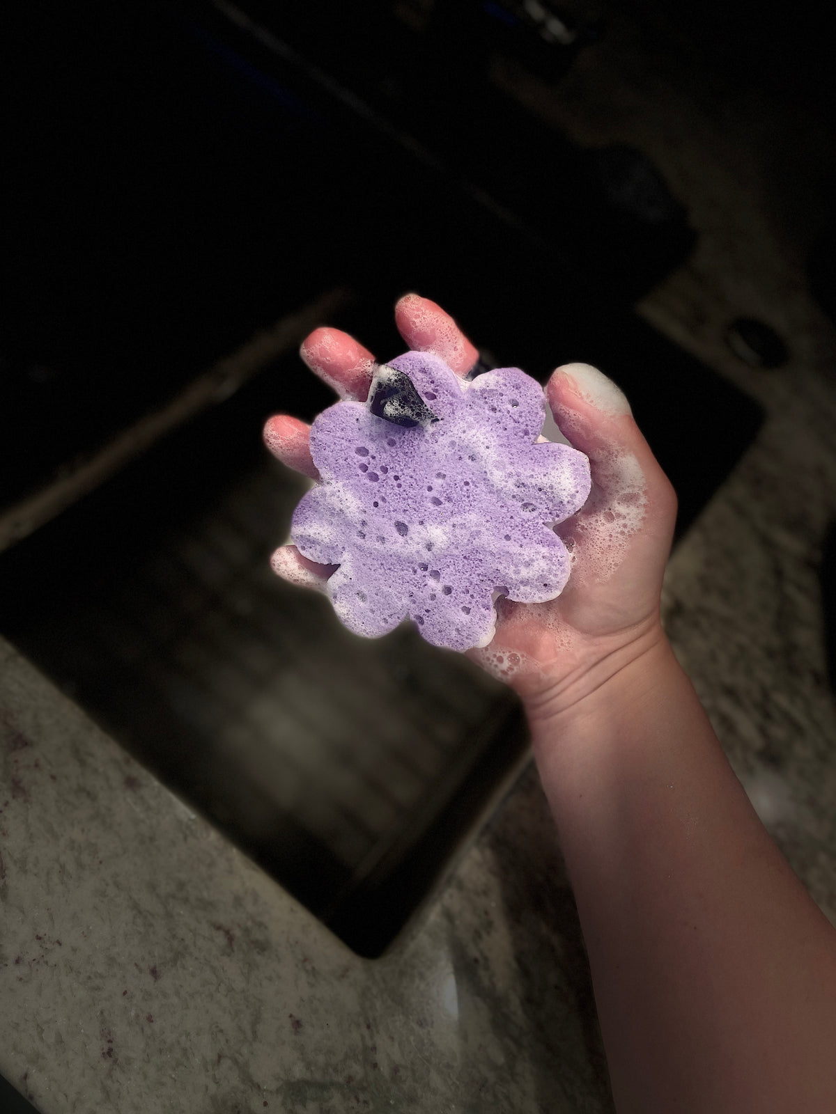 A hand holding a purple flower-shaped sponge with soap foam, set against a dark kitchen sink background.