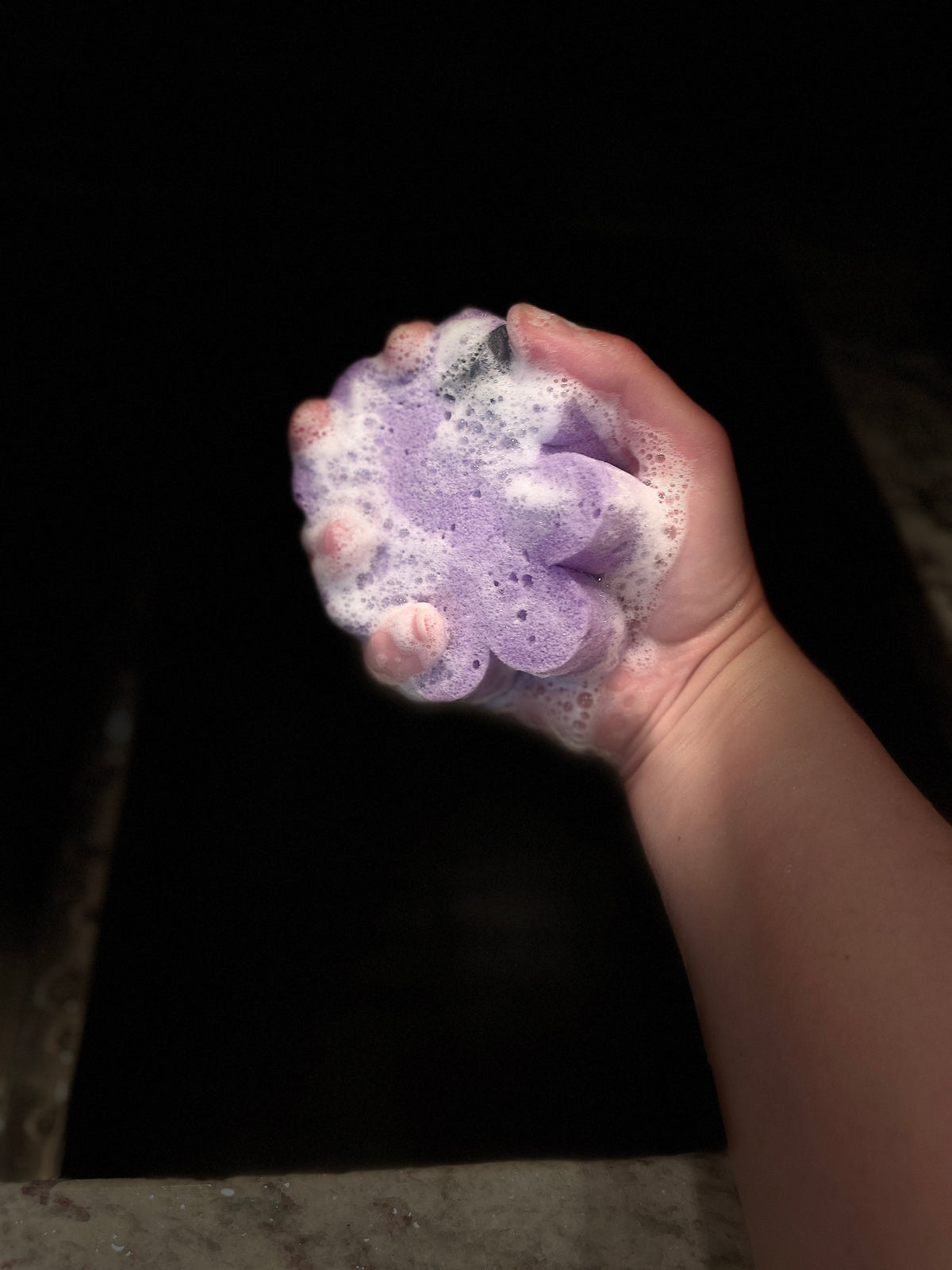 A hand holding a purple soapy sponge against a dark background.