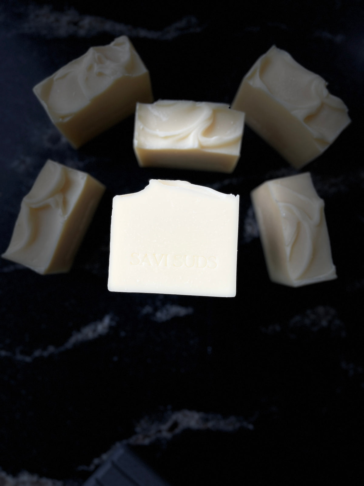 Six bars of handmade soap in a cream shade, arranged on a dark black marble surface. One bar is elevated in the center and has 'SAVI SUDS' stamped on it.