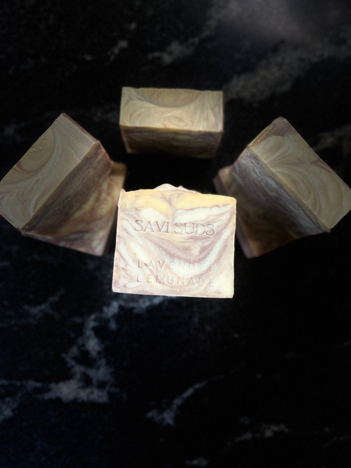 Four square bars of artisanal soap with a marbled cream, beige, and brown swirl pattern, arranged on a dark black marble surface. The center bar is upright and features the embossed text 'SAVI SUDS'