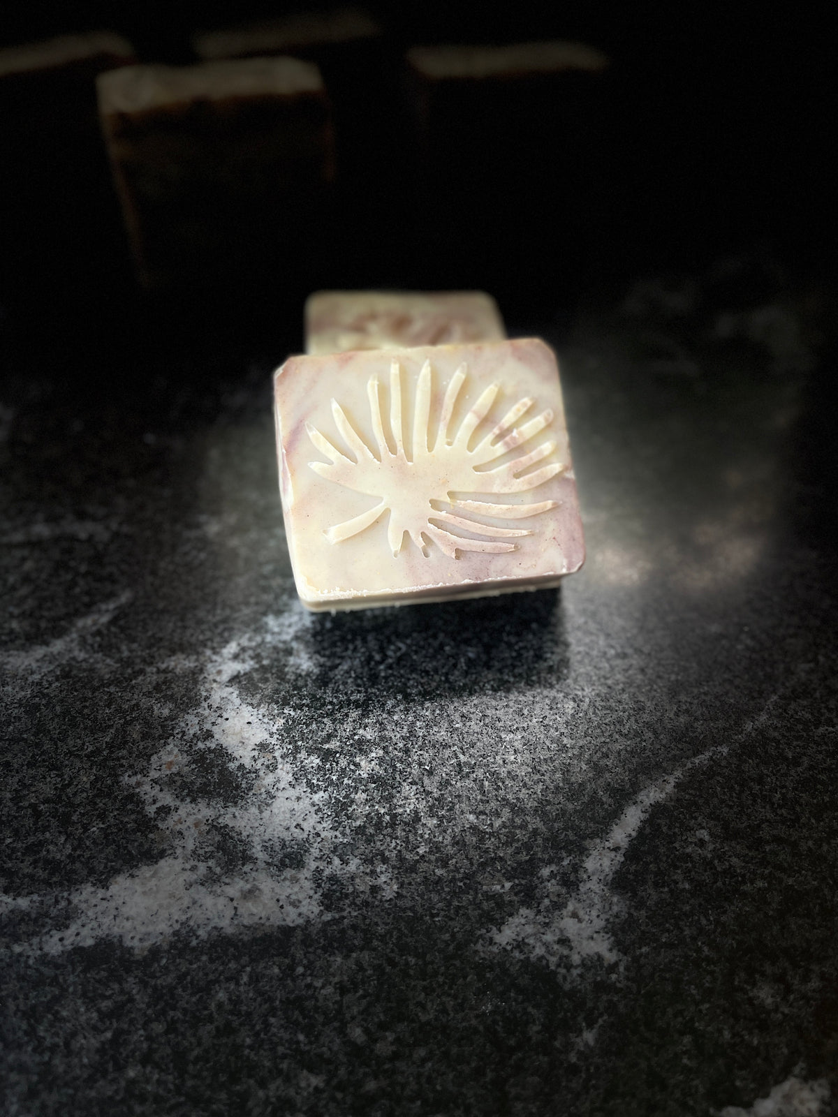 A square handmade soap bar with a pale purple and cream marbled pattern, embossed with a leaf design on one side, resting on a dark textured surface.