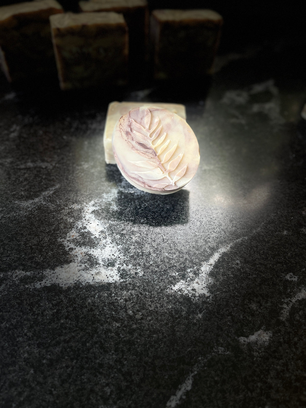 A round handmade soap bar with a pale purple and cream marbled pattern, embossed with a leaf design on one side, resting on a dark textured surface.