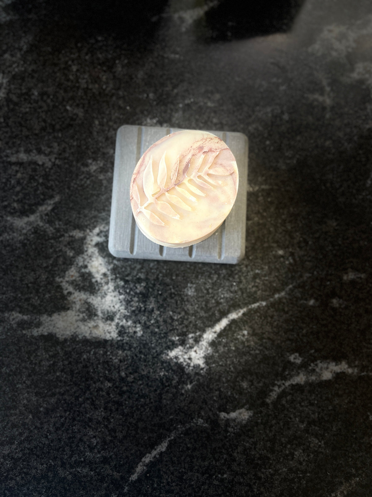 An oval-shaped pale purple marbled soap bar with a leaf vein pattern, resting on a gray square block over a dark marbled countertop.