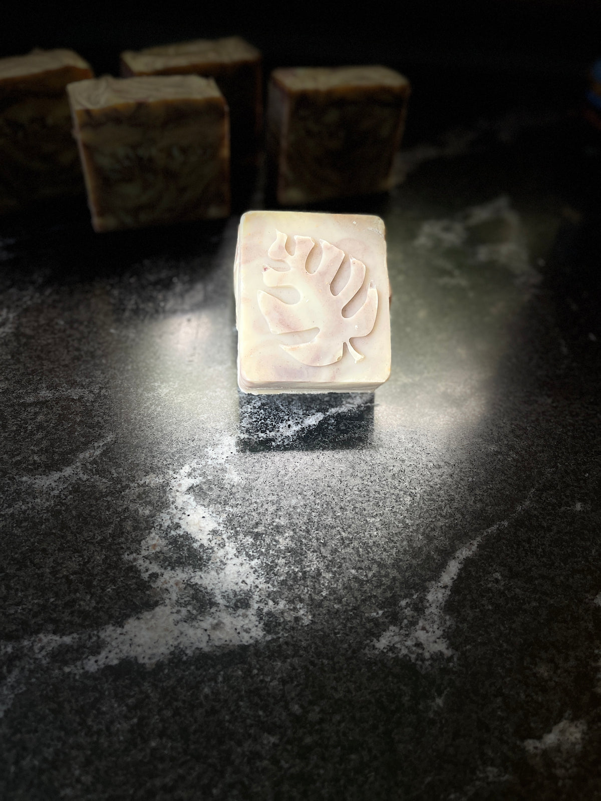 A square handmade soap bar with a molded leaf design on top, leaning on its side on a dark marble-like surface with other blurred soap bars in the background.